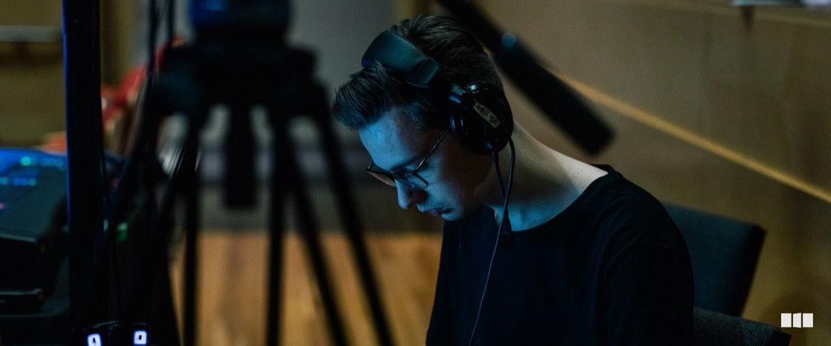 A young person wearing headphones and glasses focuses intently at a workstation bathed in blue light, with a camera tripod visible in the foreground.