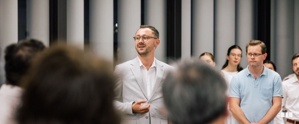 A man in a light grey blazer addresses a group of people gathered in a modern hall