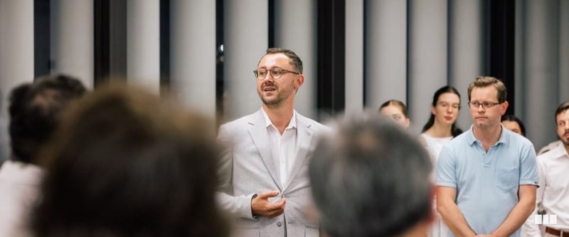 A man in a light grey blazer addresses a group of people gathered in a modern hall
