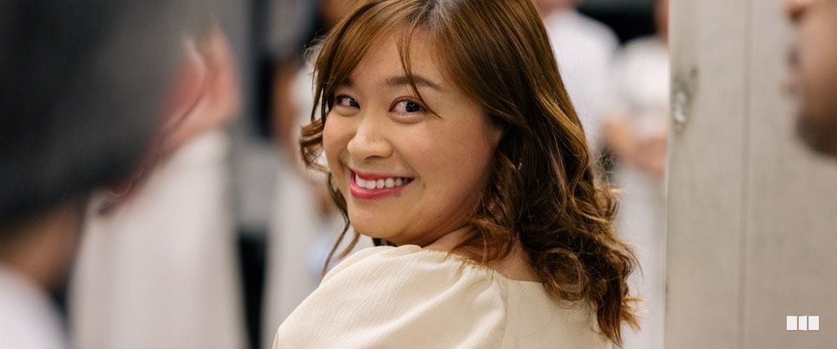 A young woman with auburn hair smiles warmly over her shoulder in a bright indoor setting among other people dressed in white.