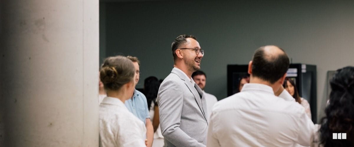 A smiling man in a light grey blazer and glasses mingles with a group of people in a casual indoor setting.