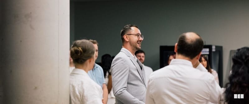 A smiling man in a light grey blazer and glasses mingles with a group of people in a casual indoor setting.