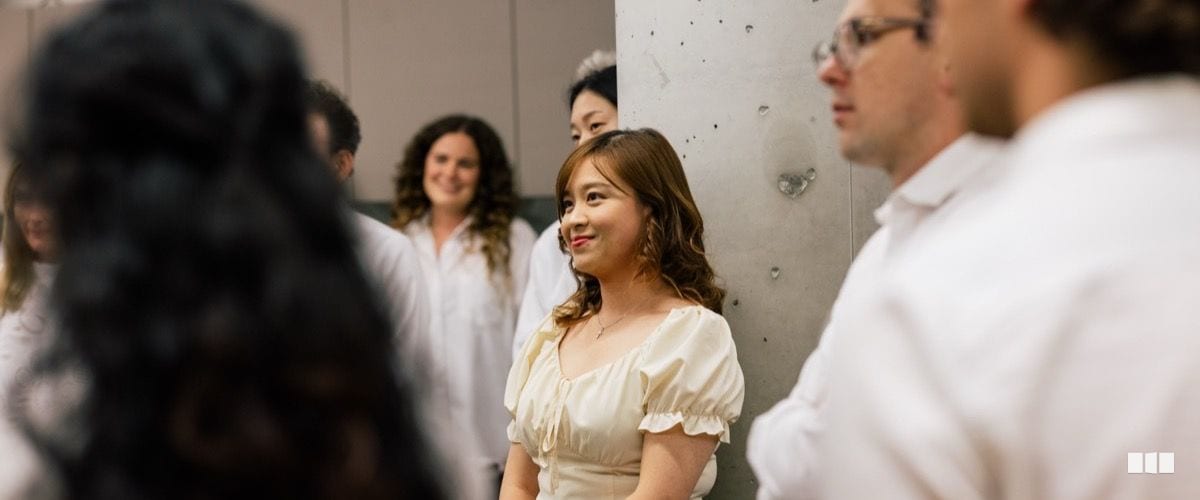 A young woman in a cream dress smiles warmly while standing with a group of people dressed in white during what appears to be a concert or rehearsal gathering.
