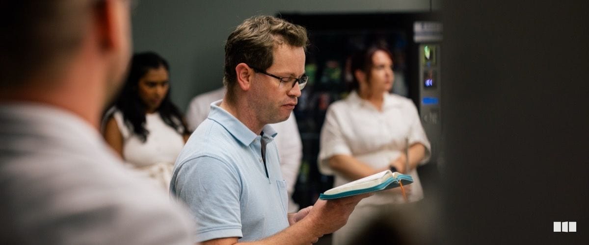A man wearing glasses reads from an open book during what appears to be a rehearsal or gathering, with other attendees visible in the background near a vending machine.