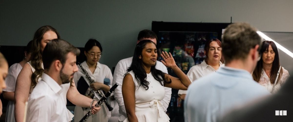 A diverse group of choir and orchestra members in white attire rehearse together, with a clarinetist in the foreground and a soloist raising her hand expressively in the centre.