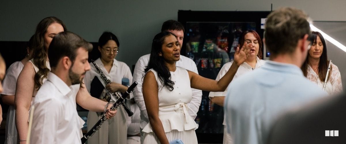 A group of musicians dressed in white, including clarinet players and a central female soloist gesturing expressively, rehearse or perform in a backstage or rehearsal room setting.