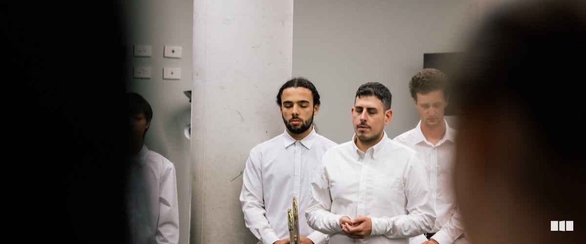 Several young male musicians in white dress shirts stand with eyes closed, one holding a brass instrument, appearing to pray or focus before a performance.
