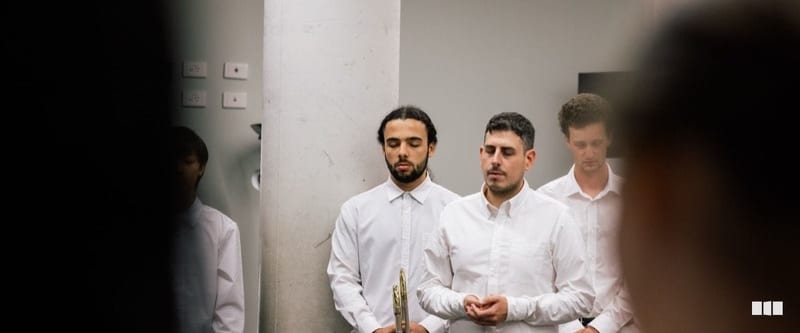 Several young male musicians in white dress shirts stand with eyes closed, one holding a brass instrument, appearing to pray or focus before a performance.