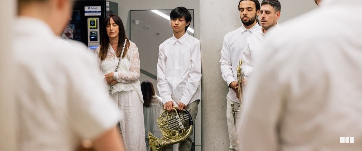 A group of musicians dressed in white stand in a backstage corridor holding a French horn and other instruments before a performance