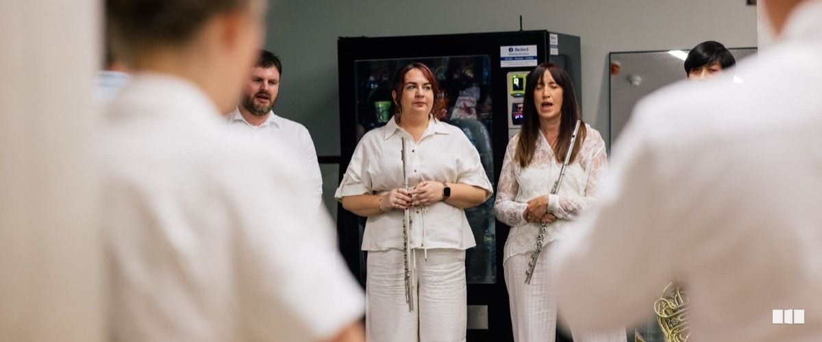 Two flautists and a male musician dressed in white gather backstage near a vending machine, appearing to rehearse or converse before a performance.