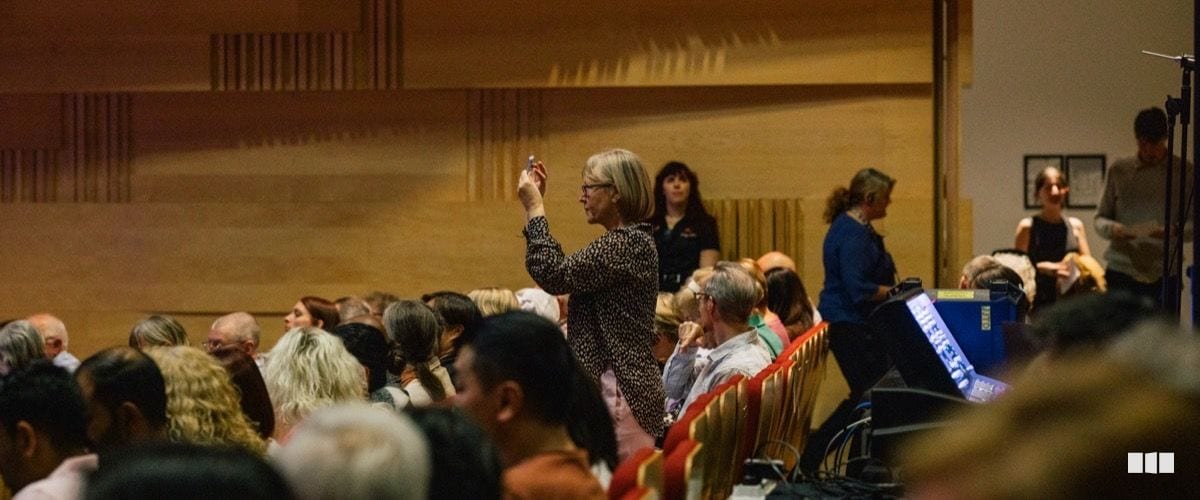 An audience member raises her hand during what appears to be a Q&A or interactive session in a warmly lit concert hall