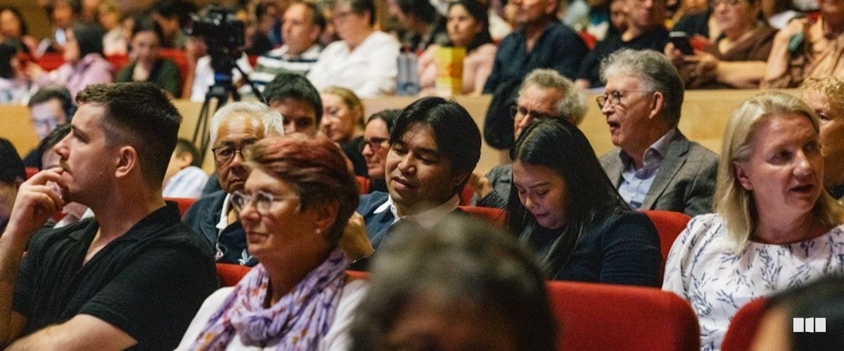A diverse audience of adults watches attentively from red theatre seats during a performance.