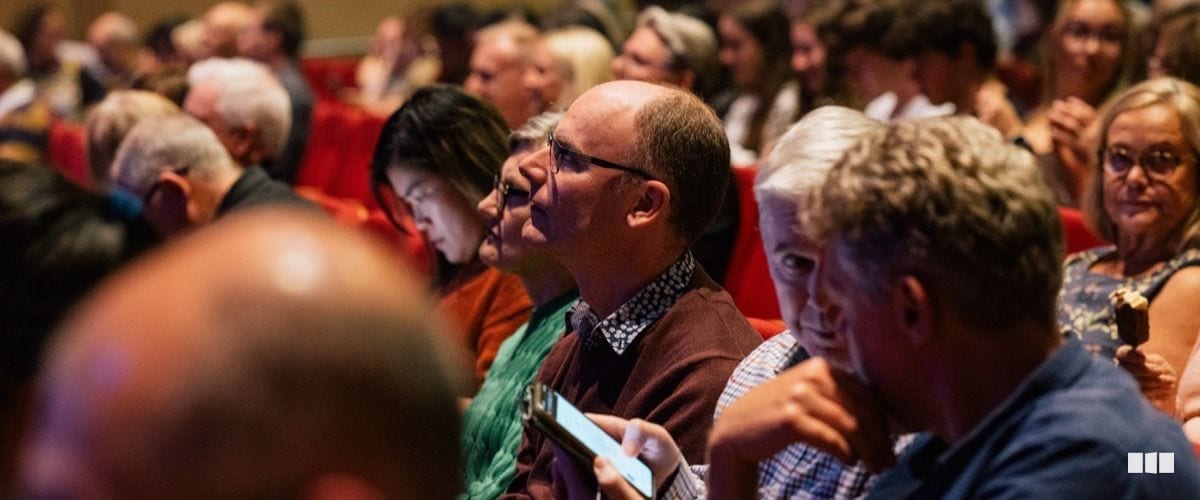 A diverse audience of adults and young people watch attentively from red theatre seats during a performance.
