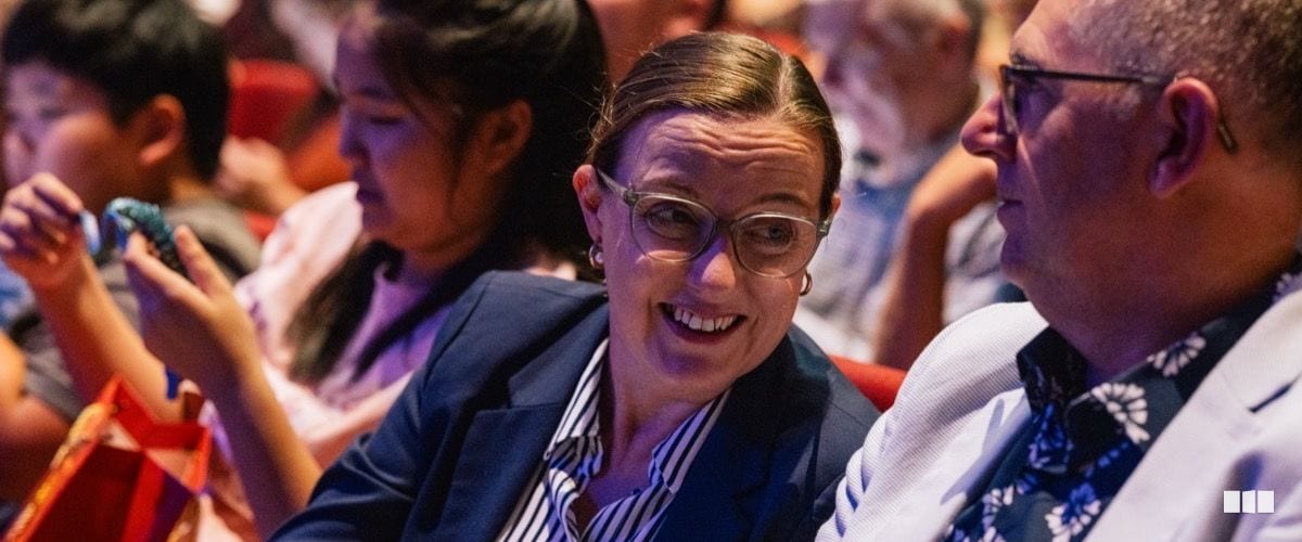 A smiling woman in glasses and a navy blazer chats with a man in a floral shirt while seated in an auditorium audience.