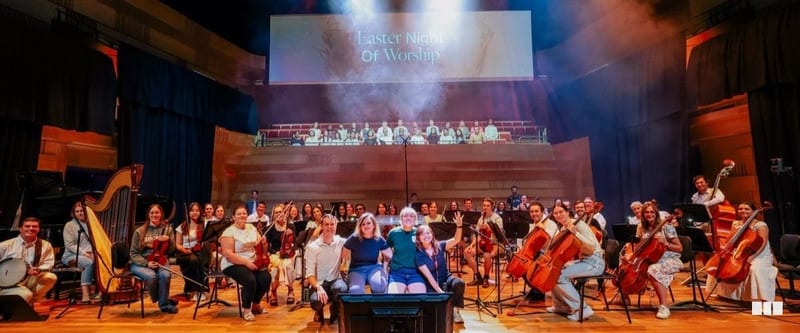 The full orchestra and choir gather on stage for a group photo at the Easter Night of Worship event in a concert hall