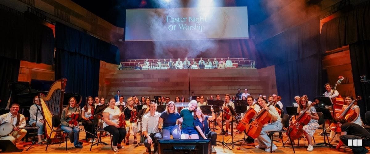 The full orchestra and choir pose together on stage after an Easter Night of Worship concert in a concert hall venue
