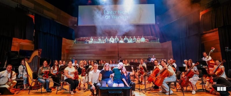 The full orchestra and choir pose together on stage after an Easter Night of Worship concert in a concert hall venue