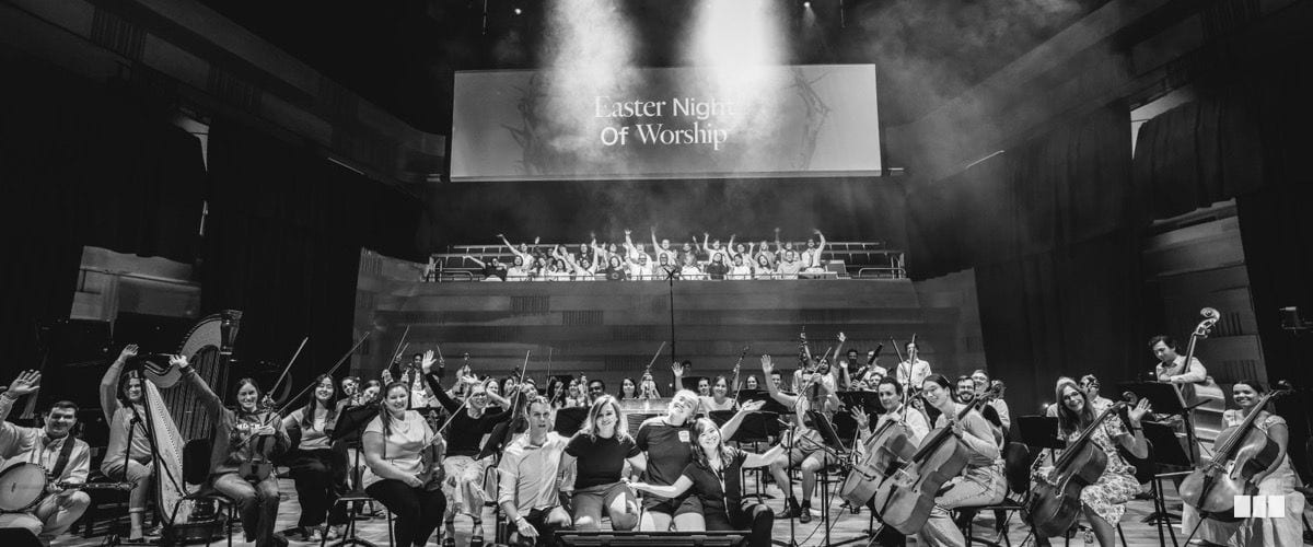 The full orchestra and choir gather on stage for a group photo at the Easter Night of Worship event, with hands raised in celebration.