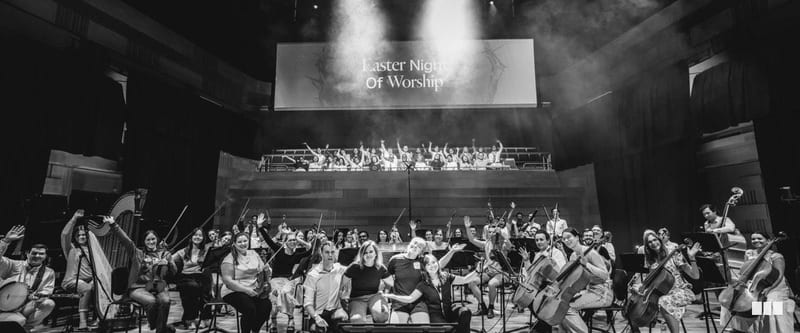 The full orchestra and choir gather on stage for a group photo at the Easter Night of Worship event, with hands raised in celebration.