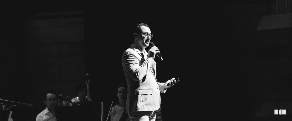 A suited man speaks into a microphone on stage before an orchestra during a concert