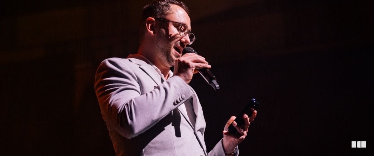 A man in a light grey suit speaks energetically into a handheld microphone on a darkened stage while holding a small device in his other hand