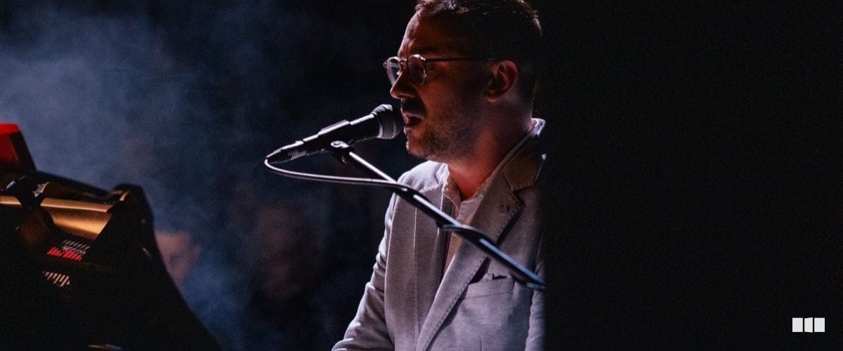 A male keyboardist wearing glasses and a white blazer performs and sings into a microphone on a dimly lit stage with atmospheric haze.