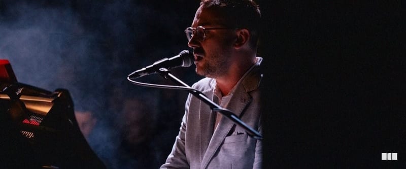 A male keyboardist wearing glasses and a white blazer performs and sings into a microphone on a dimly lit stage with atmospheric haze.