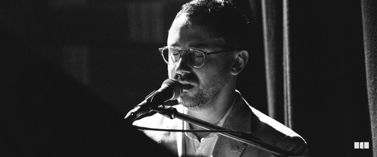 A male vocalist with glasses performs at a microphone on a dimly lit stage