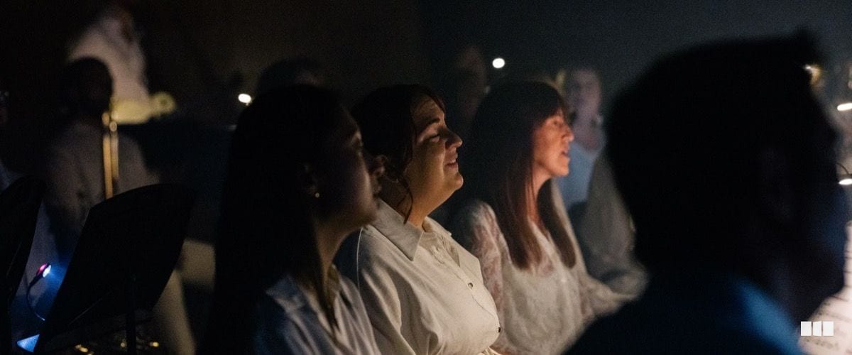Choir singers dressed in white perform with focused expressions in a dimly lit concert setting
