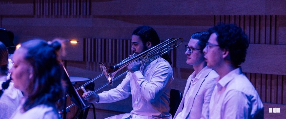 A trombone player in a white shirt performs alongside fellow musicians in a warmly lit concert hall with acoustic panelling.