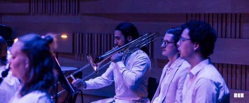 A trombone player in a white shirt performs alongside fellow musicians in a warmly lit concert hall with acoustic panelling.