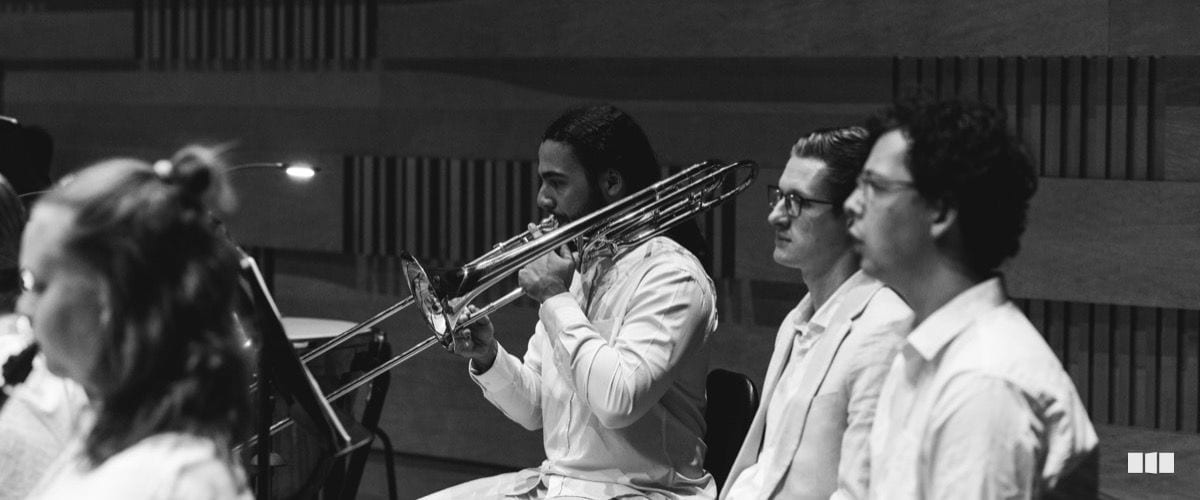 A trombonist plays during an orchestral rehearsal or performance in a concert hall, seated alongside fellow musicians in white attire.