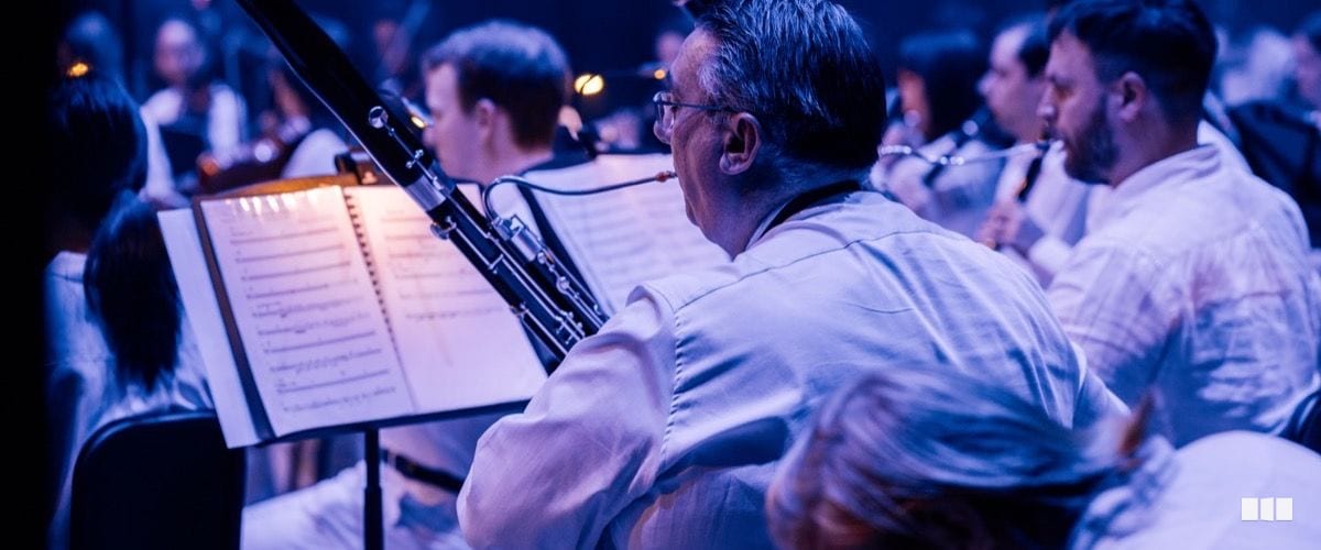 A bassoonist reads from a music stand during an orchestral performance under blue stage lighting, with fellow wind players visible behind.