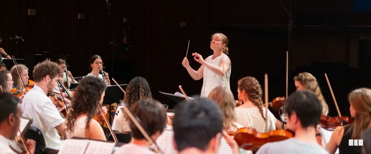 A conductor raises a baton with expressive intensity while leading a string-heavy orchestra during a rehearsal or performance.