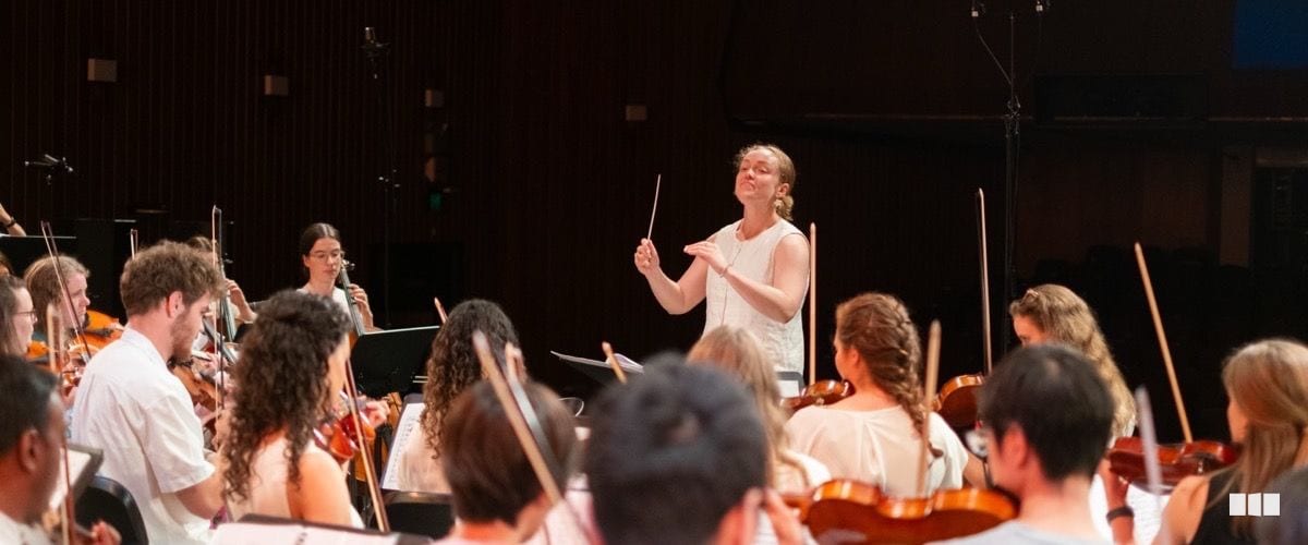 A conductor leads a string orchestra rehearsal on stage, gesturing expressively with baton raised
