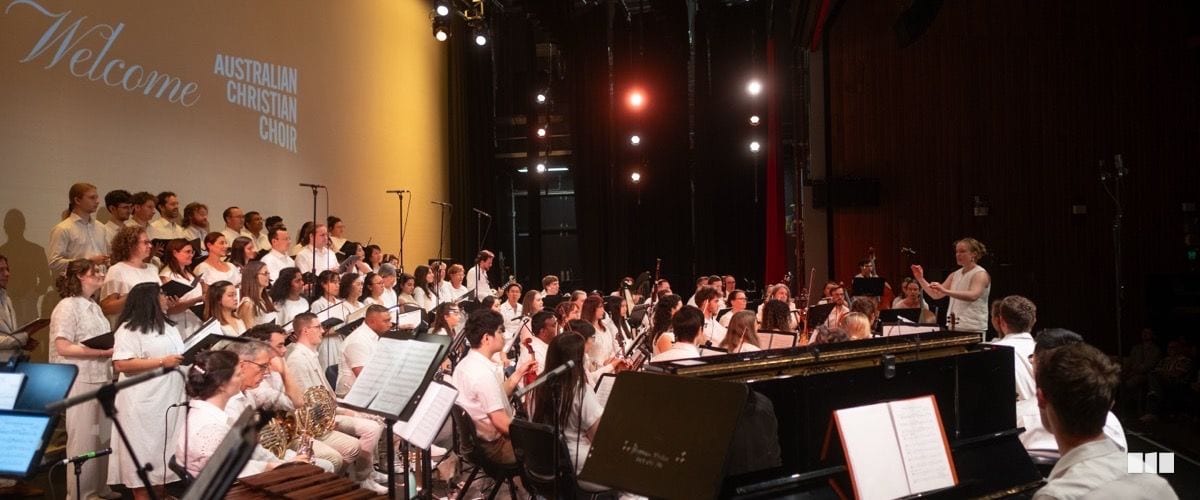 The Australian Christian Choir and orchestra perform together on stage in white attire, led by a conductor at the podium.