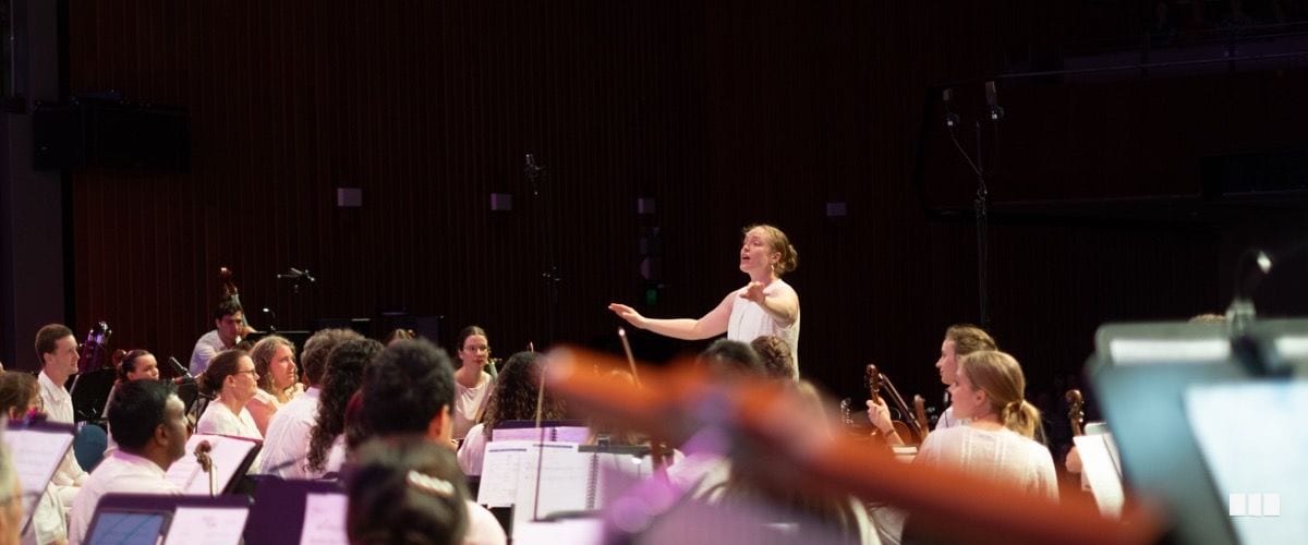 A female conductor leads a youth orchestra on stage, gesturing expressively with a baton during a performance