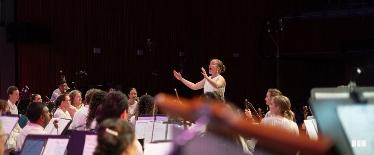 A conductor in white leads a full orchestra on stage during a concert performance, with musicians and music stands filling the foreground.