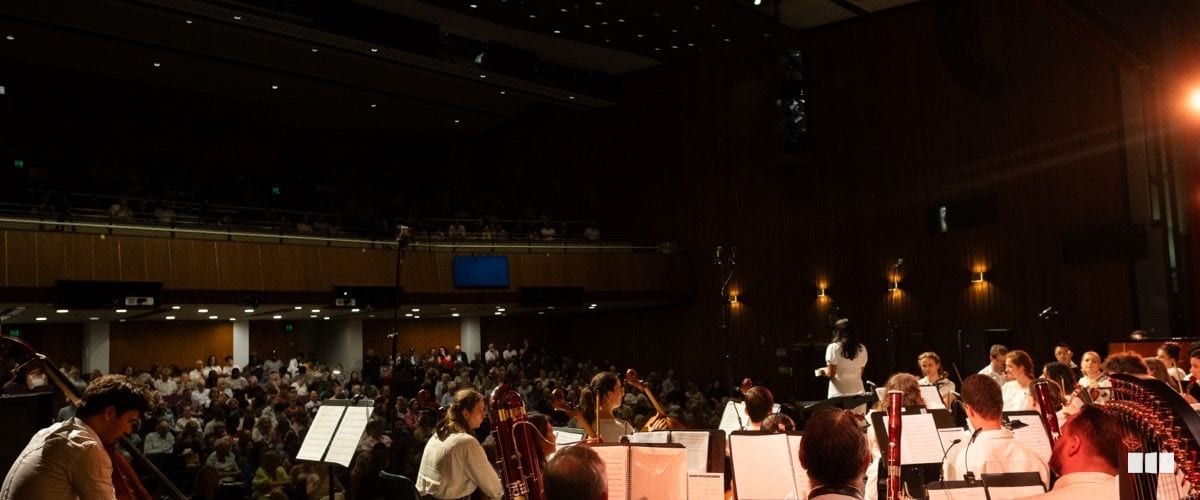 A symphony orchestra performs on stage from the conductor's perspective, facing a large seated audience in a darkened concert hall