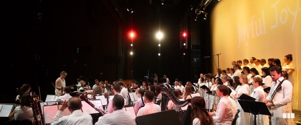 A large ensemble of orchestral musicians and choir members dressed in white perform together on a theatre stage beneath a projected 'Joyful Joy' backdrop.