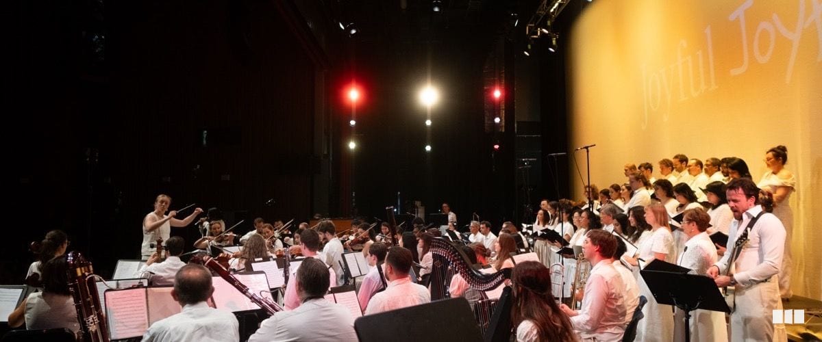 A large orchestra and choir dressed in white perform together on a warmly lit stage beneath a projected 'Joyful Joy' backdrop.