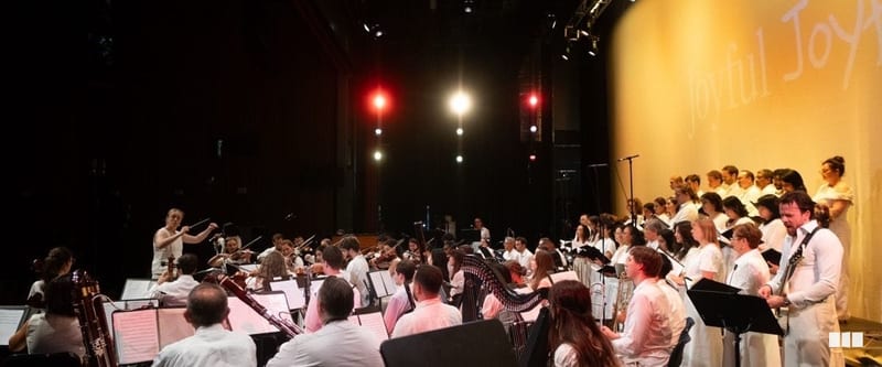 A large orchestra and choir dressed in white perform together on a warmly lit stage beneath a projected 'Joyful Joy' backdrop.
