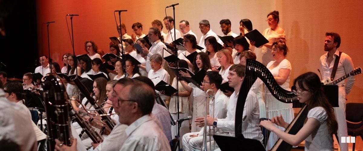 A large choir in white attire performs on stage alongside orchestral musicians including a harpist, brass players, and woodwind players in a warmly lit concert hall.