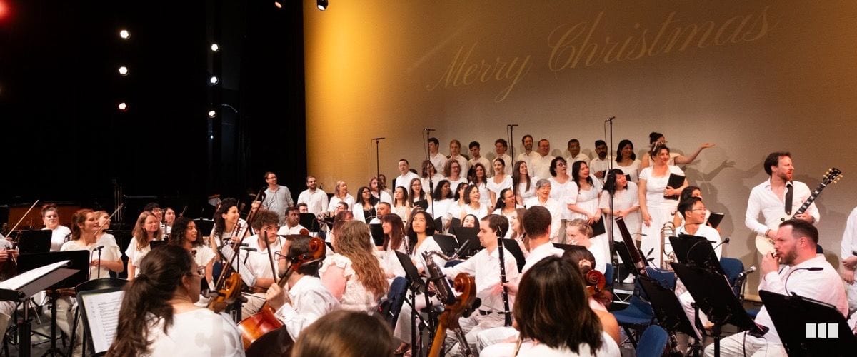 A large ensemble of orchestral musicians and choir members dressed in white perform together on stage beneath a projected 'Merry Christmas' message.